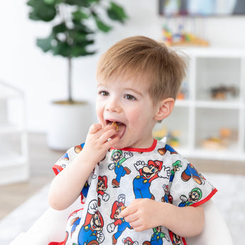A boy wearing a Bumkins Junior Bib: Super Mario™ & Luigi eats a snack in a bright room with toy filled shelves and a plant.