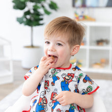 Load image into Gallery viewer, A boy wearing a Bumkins Junior Bib: Super Mario™ & Luigi eats a snack in a bright room with toy filled shelves and a plant.