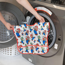 Load image into Gallery viewer, A person holds Bumkins Junior Bib: Super Mario™ & Luigi in front of an open front-loading washing machine.