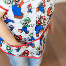 Load image into Gallery viewer, Close-up of a child wearing a Bumkins Junior Bib: Super Mario™ & Luigi with their hand in the pocket.