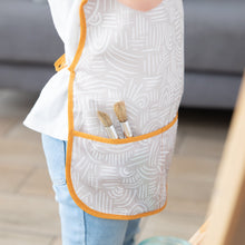 Load image into Gallery viewer, Close-up of a child in a Bumkins Short-Sleeved Smock: Wander with paint brushes in the pocket.