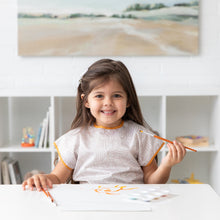 Load image into Gallery viewer, A child in a Bumkins Short-Sleeved Smock: Wander smiles, holding a paintbrush at a white table in a sunlit room.