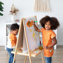 Load image into Gallery viewer, Two kids paint at an easel in a bright room; one smiles, wearing a Bumkins Short-Sleeved Smock: Grounded, while the other focuses.