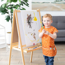 Load image into Gallery viewer, A child in a Bumkins Short-Sleeved Smock: Grounded holds a paintbrush by an easel with art, with a large plant and couch in the background.