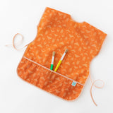 Bumkins Short-Sleeved Smock: Grounded holds paintbrushes in its pocket while laying on a white background.