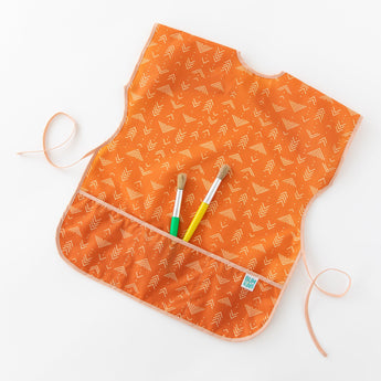 Bumkins Short-Sleeved Smock: Grounded holds paintbrushes in its pocket while laying on a white background.