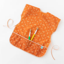 Load image into Gallery viewer, Bumkins Short-Sleeved Smock: Grounded holds paintbrushes in its pocket while laying on a white background.