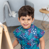 A child beams at the camera in a Bumkins Short-Sleeved Smock: Animal Prints, beside an easel, gray chair, and wooden table.