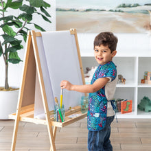 Load image into Gallery viewer, A young boy in a Bumkins Short-Sleeved Smock: Animal Prints paints on an easel in a bright room with a large plant and shelves as a backdrop.