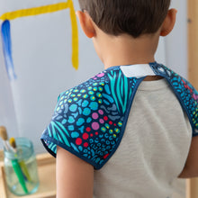 Load image into Gallery viewer, A child in a Bumkins Short-Sleeved Smock: Animal Prints stands by an easel with brushes nearby, with the hook-and-loop neck closure visible.