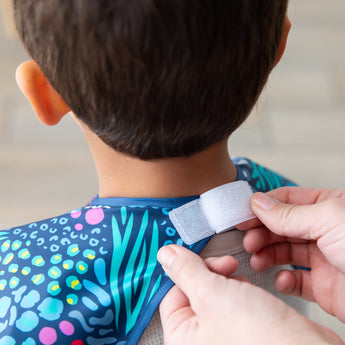 Hands fastens the hook-and-loop closure of a Bumkins Short-Sleeved Smock: Animal Prints around a child's neck.