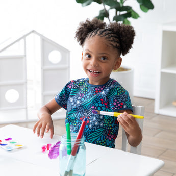 A child paints happily at a white table, wearing a Bumkins Short-Sleeved Smock: Animal Prints in a bright, airy room.