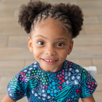 A smiling girl with braided hair in a Bumkins Short-Sleeved Smock: Animal Prints sits on a chair against a wooden floor.