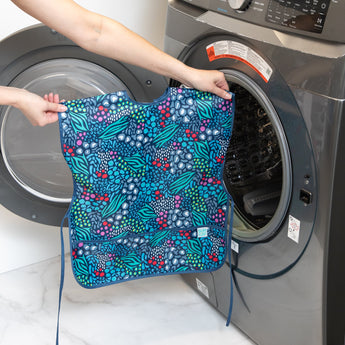 A person holds a Bumkins Short-Sleeved Smock: Animal Prints in front of an open washing machine.
