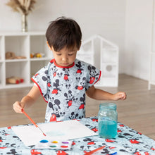Load image into Gallery viewer, A child paints at a table wearing a Bumkins Short-Sleeved Smock: Mickey Mouse Classic, with a palette and a jar of water nearby.
