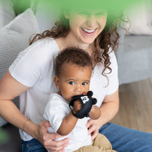 Load image into Gallery viewer, A woman smiles as her baby enjoys a Bumkins Silicone Un-Droppable Mitt Teether: Mickey Mouse on the couch, surrounded by green leaves.