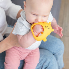 Load image into Gallery viewer, A baby with grey top and pink pants chews on a Bumkins Silicone Un-Droppable Mitt Teether: Winnie the Pooh while on an adults lap.