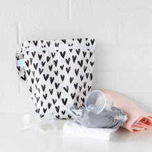 Load image into Gallery viewer, Bumkins Wet Bag: Hearts sits on a white table against a white brick wall, with cloth diapers and breast pump parts in front.