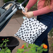 Load image into Gallery viewer, A person attaches a Bumkins Wet Bag: Hearts to a stroller using the strap, while vibrant flowers bloom in the foreground.