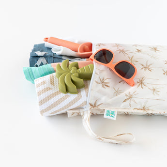 Bumkins Wet Bag: Palm Daze holds sunglasses, a toy palm tree, and beach items against a white background.