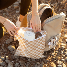 Load image into Gallery viewer, A person opens a Roamio by Bumkins Wet Bag: Sand Check next to a backpack on rocky ground, revealing a towel and water bottle inside.