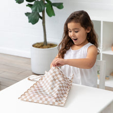 Load image into Gallery viewer, A young girl peers into a Roamio by Bumkins Wet Bag: Sand Check at a white table, with a plant in the background.