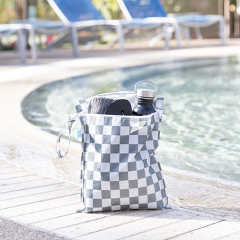 Bumkins Wet Bag: Charcoal Check holds a towel, water bottle, and flip flops poolside with empty lounge chairs in the background.