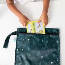 Load image into Gallery viewer, A child slips a book into a Bumkins Wet Bag: Starry Cactus, featuring cactus and moon designs, on a white table.