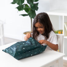 Load image into Gallery viewer, A child opens a Bumkins Wet Bag: Starry Cactus at a white table with a plant and shelves in the background.