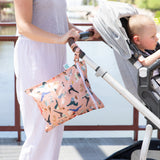 A person in white pants pushes a stroller with a baby and a Bumkins Wet Bag: Yoga Pals hanging on the handle.