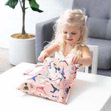 Load image into Gallery viewer, A blonde girl smiles as she opens a Bumkins Wet Bag: Yoga Pals at a white table in front of a gray couch and plant.