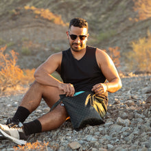 Load image into Gallery viewer, A man wearing sunglasses sits on rocky ground in golden sunlight, reaching into a Roamio by Bumkins Wet Bag: Rich Black.