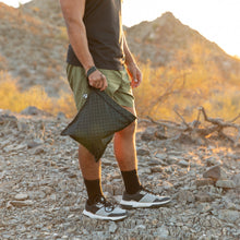 Load image into Gallery viewer, A man stands on rocky ground holding a Roamio by Bumkins Wet Bag: Rich Black with mountains in the background.