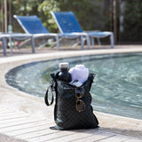 Roamio by Bumkins Wet Bag: Rich Black holds a towel, water bottle, and sunglasses poolside with empty blue lounge chairs behind.