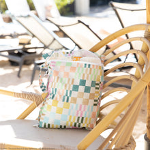 Load image into Gallery viewer, Roamio by Bumkins Wet Bag: Motley Check rests on a rattan chair poolside, holding a towel and sunscreen, with sun loungers nearby.