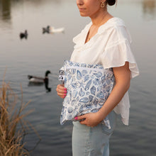 Load image into Gallery viewer, A woman in a white blouse and jeans carries a Roamio by Bumkins Wet Bag: Shells by water with ducks and reeds.