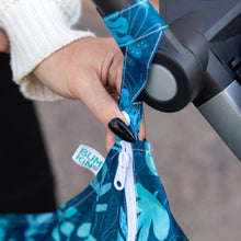 Load image into Gallery viewer, Close-up of a hand with black nails attaching a Bumkins Wet Bag: Blue Tropic to a stroller handle using the snap button on the strap.