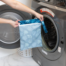Load image into Gallery viewer, A person holds Bumkins Wet Bag: Blue Tropic in front of an open front-loading washing machine.