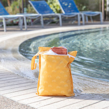 Load image into Gallery viewer, Bumkins Wet Bag: Sunshine holds clothes poolside with empty blue lounge chairs in the background.