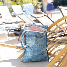 Load image into Gallery viewer, Roamio by Bumkins Wet Bag: Vintage Denim holds clothes and a water bottle on a rattan lounge chair with people relaxing in the background.