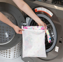 Load image into Gallery viewer, A person holds Bumkins Wet Bag: Watercolor in front of an open front-loading washing machine.