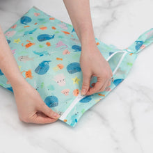 Load image into Gallery viewer, Hands zipping a Bumkins Wet Bag: Ocean Life on a marble surface.