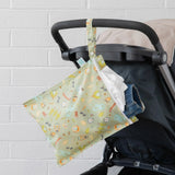 A stroller in front of a white wall with a Bumkins Wet Bag: Camp Gear hanging from the handle, filled with a white shirt and denim shorts.