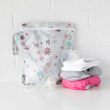 Load image into Gallery viewer, Bumkins Wet Bag: Floral sits on a white table against a white brick wall with cloth diapers and breast pump parts in front.