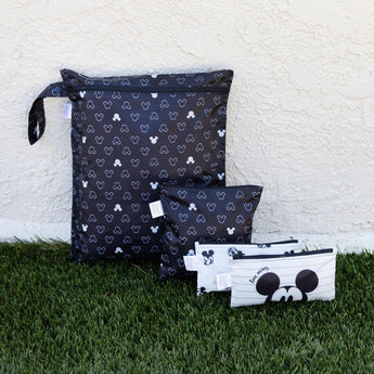 Bumkins Wet Bag: Mickey Mouse Icon Black + White sits on grass against a beige textured wall with three other bags.