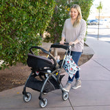A smiling woman in jeans pushes a stroller with a Bumkins Wet Bag: Mickey Mouse Classic attached to the handle on a sidewalk past greenery.