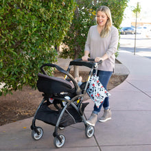 Load image into Gallery viewer, A smiling woman in jeans pushes a stroller with a Bumkins Wet Bag: Mickey Mouse Classic attached to the handle on a sidewalk past greenery.