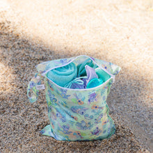 Load image into Gallery viewer, Bumkins Wet Bag: Aloha Stitch holds a towel and swimwear on a sandy beach shore.
