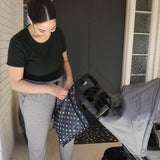 A woman in green top and gray pants zips a Bumkins Wet Bag: Super Mario™ Lineup attached to a stroller in front of a door.
