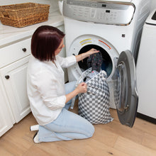 Load image into Gallery viewer, A woman kneels by a front-loading washer, placing clothes from a Bumkins Extra Large Wet Bag: Charcoal Check inside.